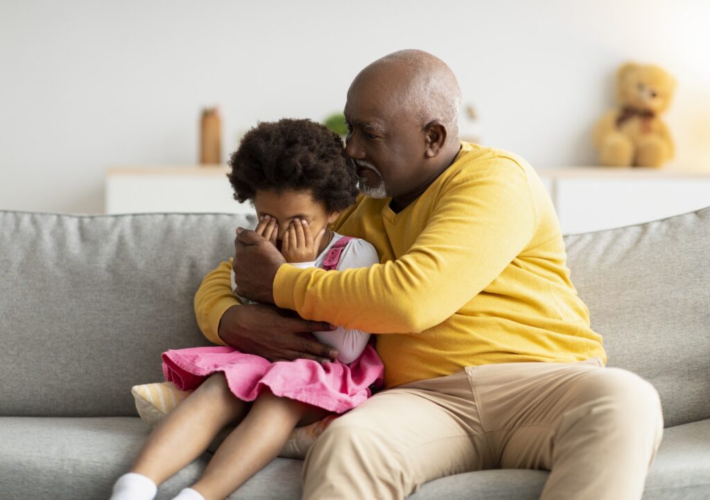 Black elderly man calming upset sad crying little girl in living room interior